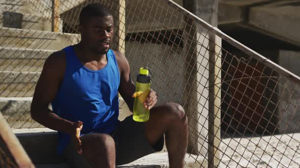 Tired african american man sitting, holding water bottle, taking break in exercise outdoors