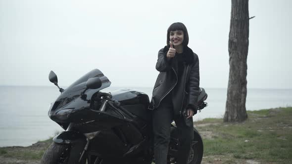 Camera Approaches To Smiling Confident Woman Standing at Motorbike and Showing Thumb Up. Portrait of