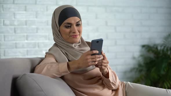 Modern Muslim Female Typing Message on Smartphone, Sitting on Home Sofa, Gadget