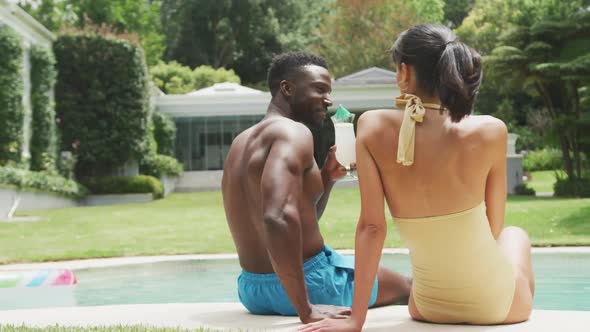 Happy diverse couple wearing swimming suits and holding hands at swimming pool in garden