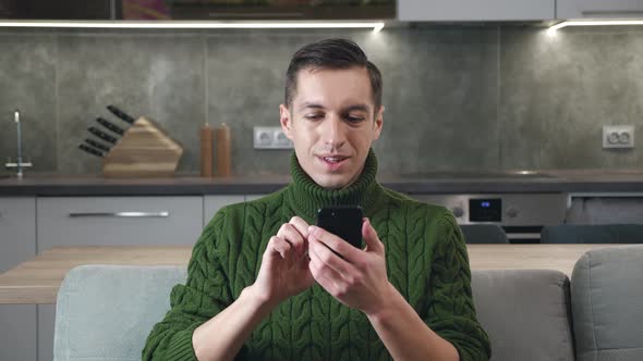 Handsome Caucasian Man Sitting on Sofa at Cosy Home and Using Smartphone Surfing Internet Social