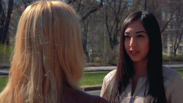 A Young Asian Woman and a Young Caucasian Woman Talk in a Street in an Urban Area