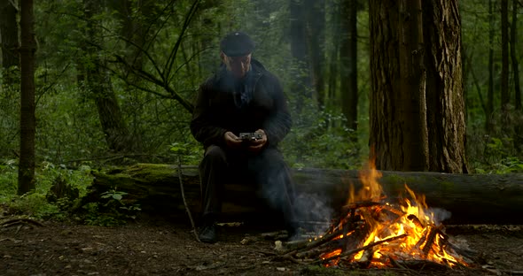 Aged Man Sits on Fallen Tree Trunk at Bonfire with Camera