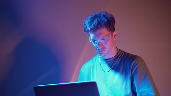 Lonely Stylish Man in Glasses Sits Working at a Laptop in Neon Lighting