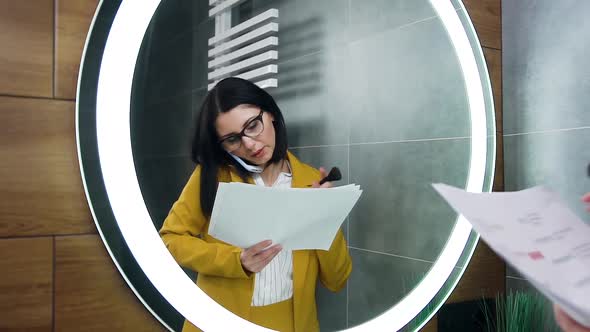 Young Business Woman Standing in Front of Mirror, Watching Documents while Talking on Phone