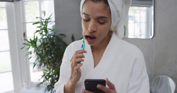 Mixed race woman brushing her teeth and using her smartphone in bathroom