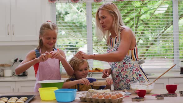 Happy caucasian mother in kitchen with daughter and son, wearing aprons baking cookies together