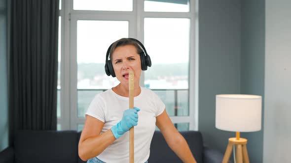 Woman in Headphones Cleans the House and Have Fun Singing with a Broom Like a Star at a Concert