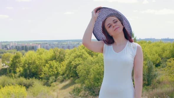 Young female in white dress and elegant hat standing in hilly terrain.