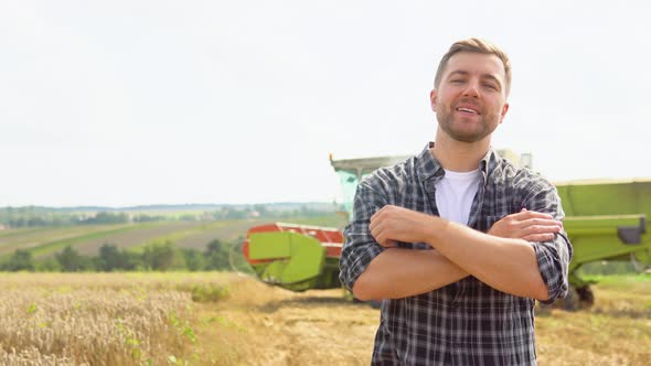 Happy Young Farmer Standing on Wheat Field While Combine Harvester in Background