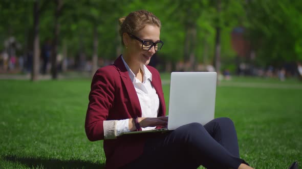 Beautiful Businesswoman with Laptop Sitting on Green Lawn Outdoors