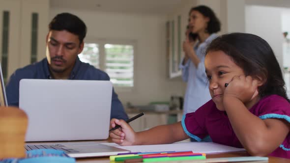 Hispanic girl sitting at table drawing with parents working in background