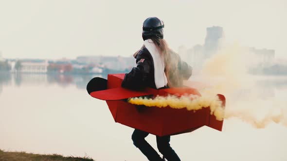 Side View of Happy Little Girl Running Along Sunset City Lake Scenery in Fun Plane Costume with