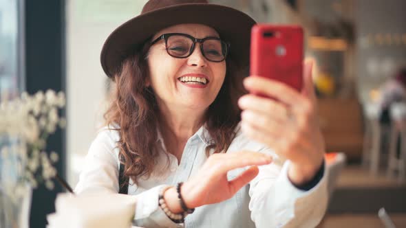 A Mature Stylish Woman in Hat and Glasses Taking a Video Call on a Smartphone