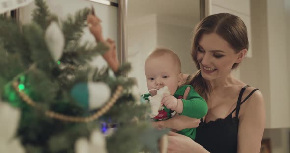 Happy Mother Holds Her Baby and Entertains Him with Toys on the Christmas Tree