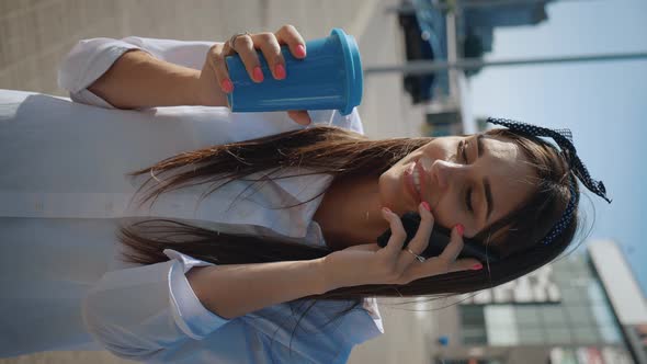 Vertical Video of Young Woman Talking on the Phone and Drinking Coffee on Background of Office