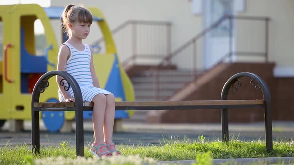 Sad Child Girl Waiting for Her Mother Sitting on a Bench on Summer Playground in Kindergarten Yard
