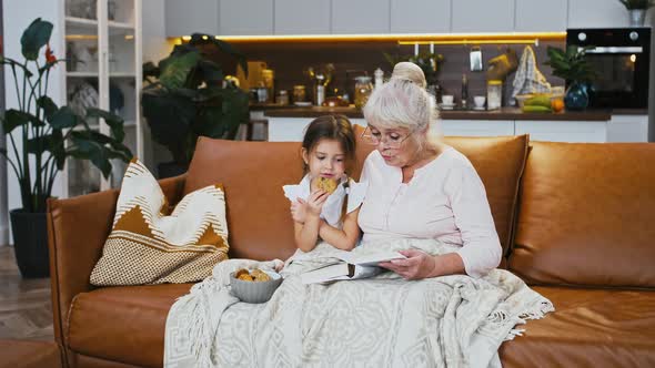 Grandmother Reading Book for Little Granddaughter Who Eating Cookies Sitting on Comfortable Sofa