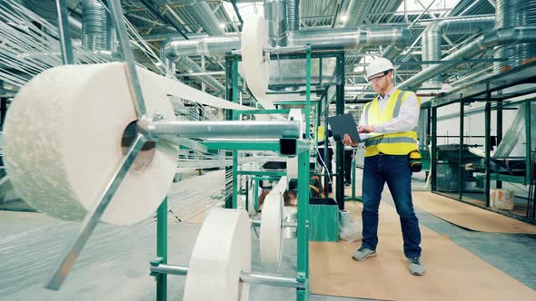 Factory Worker is Observing Rolls of Fiberglass and Using a Laptop