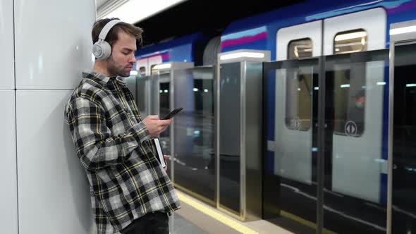 Young Guy Waits for a Train in the Subway Uses a Smartphone and Listens to Music with Wireless