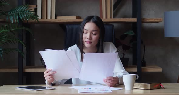 Asian Woman Indoor Sitting in the Workplace While Looking Through Paper Documents in Loft Apartment