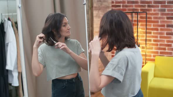 Cute Woman Closing Eyes Cuts Off a Strand of Hair with Scissors in Front of a Mirror