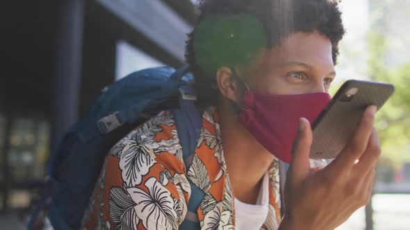 African american man in city, wearing face mask sitting using smartphone in street