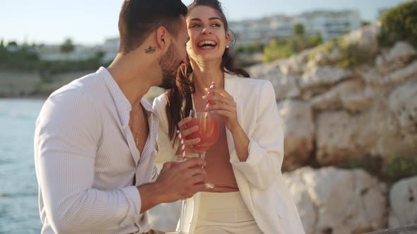 Smiling Hispanic Couple with Cocktails Kissing Against Sea