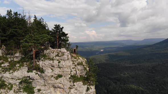 A Young Man in a Black T-shirt and Shorts Stands on the Edge of a High Cliff and Looks Around