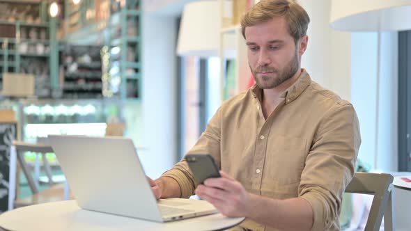 Young Man Using Smartphone and Laptop in Cafe