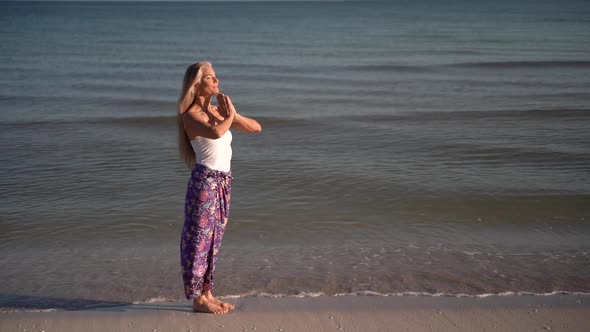 Slow motion of mature woman in sarong in a prayer pose on a beach at sunrise or sunset.