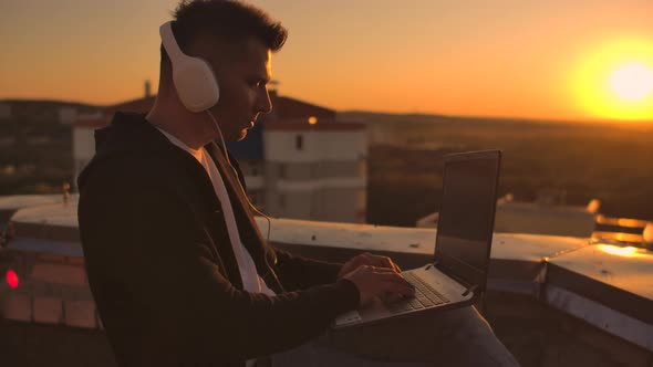 A Man Freelancer in Headphones Standing on the Roof at Sunset Writes on the Keyboard Code Pages