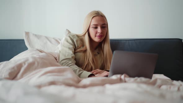 Smiling blond woman working on laptop and looking to the side in bed