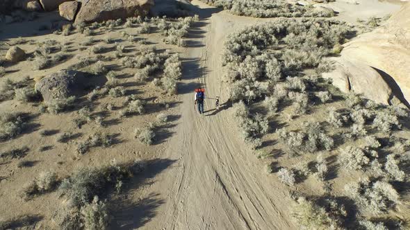 Aerial shot of a young man backpacking with his dog on a dirt road in a mountainous desert.