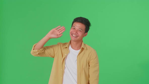 The Happy Young Asian Man Waving Hand And Say Bye Bye While Standing On Green Screen In The Studio