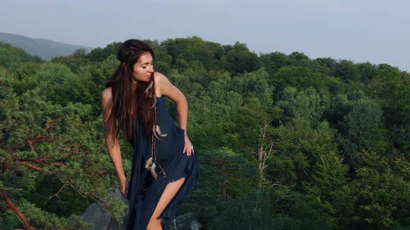 Beautiful Wild Medieval Woman in a Blue Dress Interwoven Feathers in Hair Stands Against of Forest