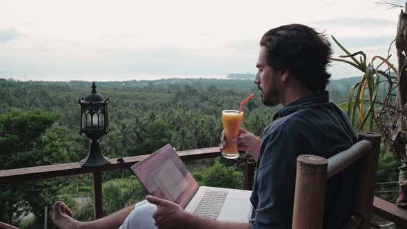 A young, handsome man with a beard works on a laptop in a cafe overlooking an incredible sunset.