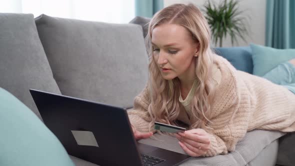 Beautiful Blonde Lying on the Couch Makes Online Payments with a Bank Card Using a Laptop at Home