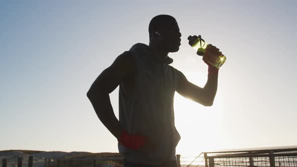 African american man drinking water, taking break in exercise outdoors by the sea