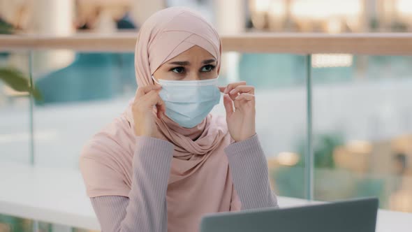 Young Arab Businesswoman in Hijab Works at Laptop Looks Around Muslim Girl Lowers Medical Mask