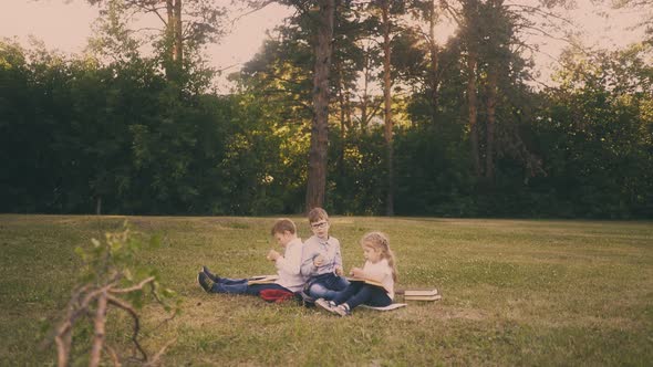 Schoolboys with Little Girl Have Lunch on Grass in Park