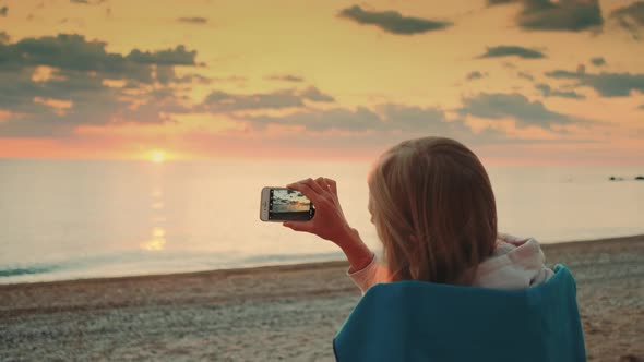 Young Woman Making Video of Sunset with Smartphone Sitting on the Seashore