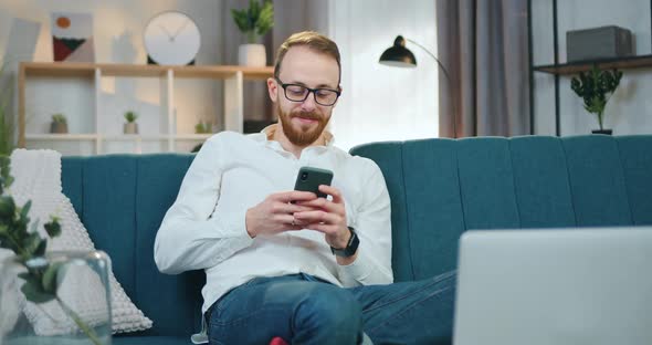 Man in Glasses Sitting in Relaxed Pose on Soft Couch at Home and Browsing Phone Apps