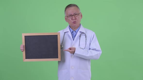 Stressed Mature Japanese Man Doctor Holding Blackboard and Giving Thumbs Down