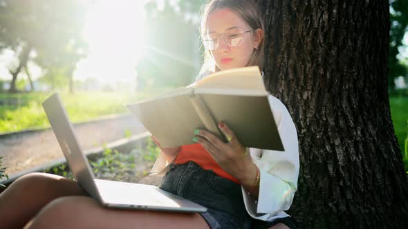 Beautiful Teenage Girl Sitting in Park with Laptop and Reading Book Aloud