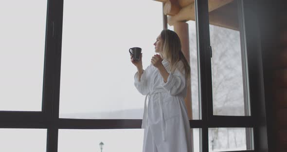 Young Beautiful Woman in White Bathrobe Drink Coffee By the Window at Home in the Morning