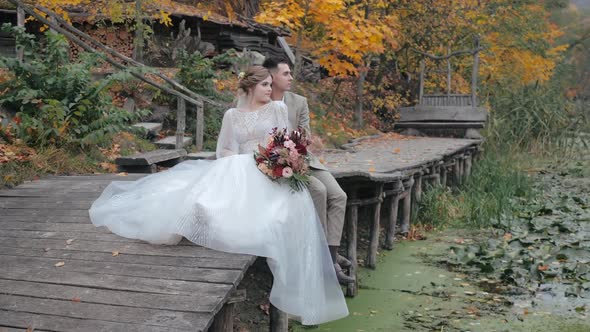 Beautiful Bride in Wedding Dress and Bouquet of Flowers Sits on a Wooden Bridge, Behind Her Hugs the