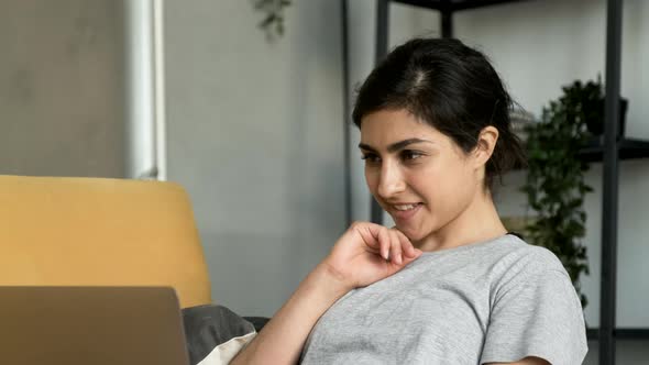 Happy young adult Indian woman sitting in cozy living room on couch holding using laptop