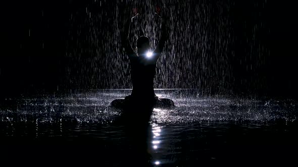 Black Silhouette Looms Under the Streams of Rain with Folded Hands in a Lotus Position. Studio Light