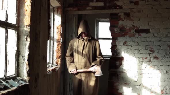 a Man in a Long Cloak with a Hood Walks in an Abandoned Building with Bloody Ax